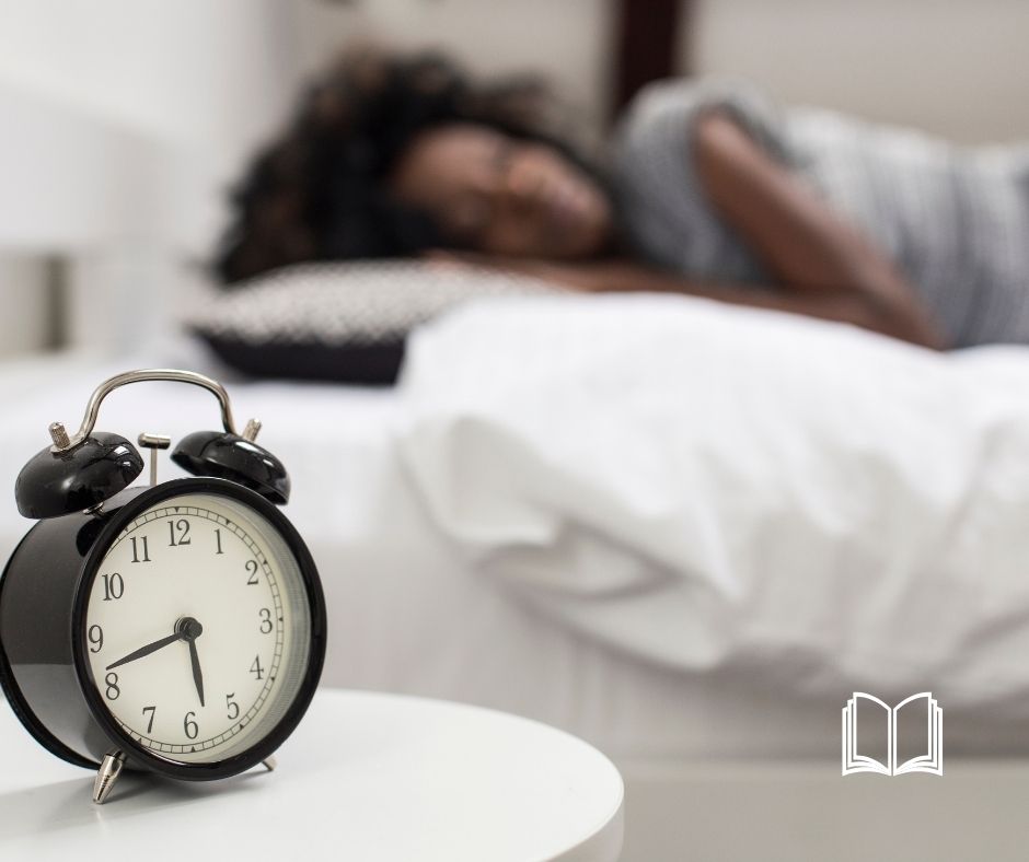 Woman sleeping in the background with a classic alarm clock in the foreground.