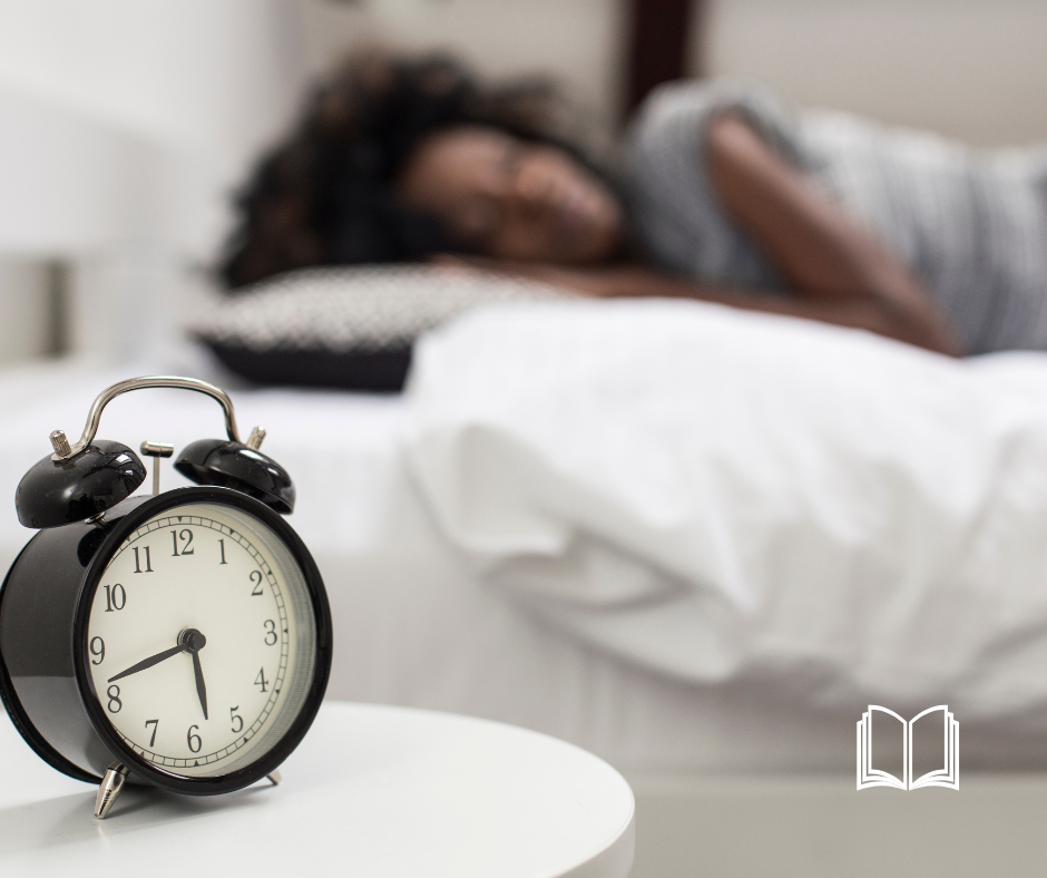 Woman sleeping in the background, and the foreground a classic style alarm clock.