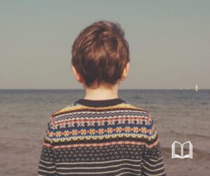 Back of a little boy in a sweater who is facing the ocean.
